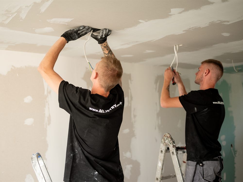 Dwóch instalatorów wpina przewody w punkty oświetleniowe w suficie remontowanego mieszkania