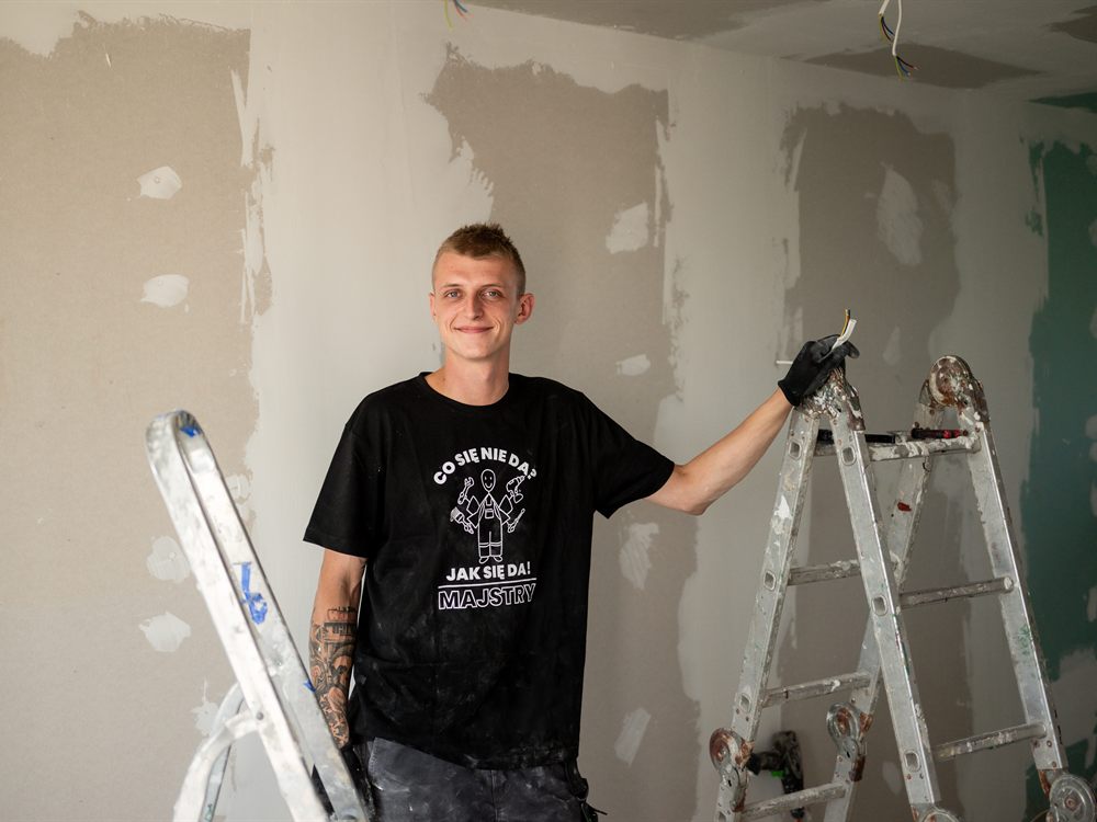 Szymon Jaciński w remontowanym mieszkaniu, między dwiema drabinami, z przewodem w dłoni